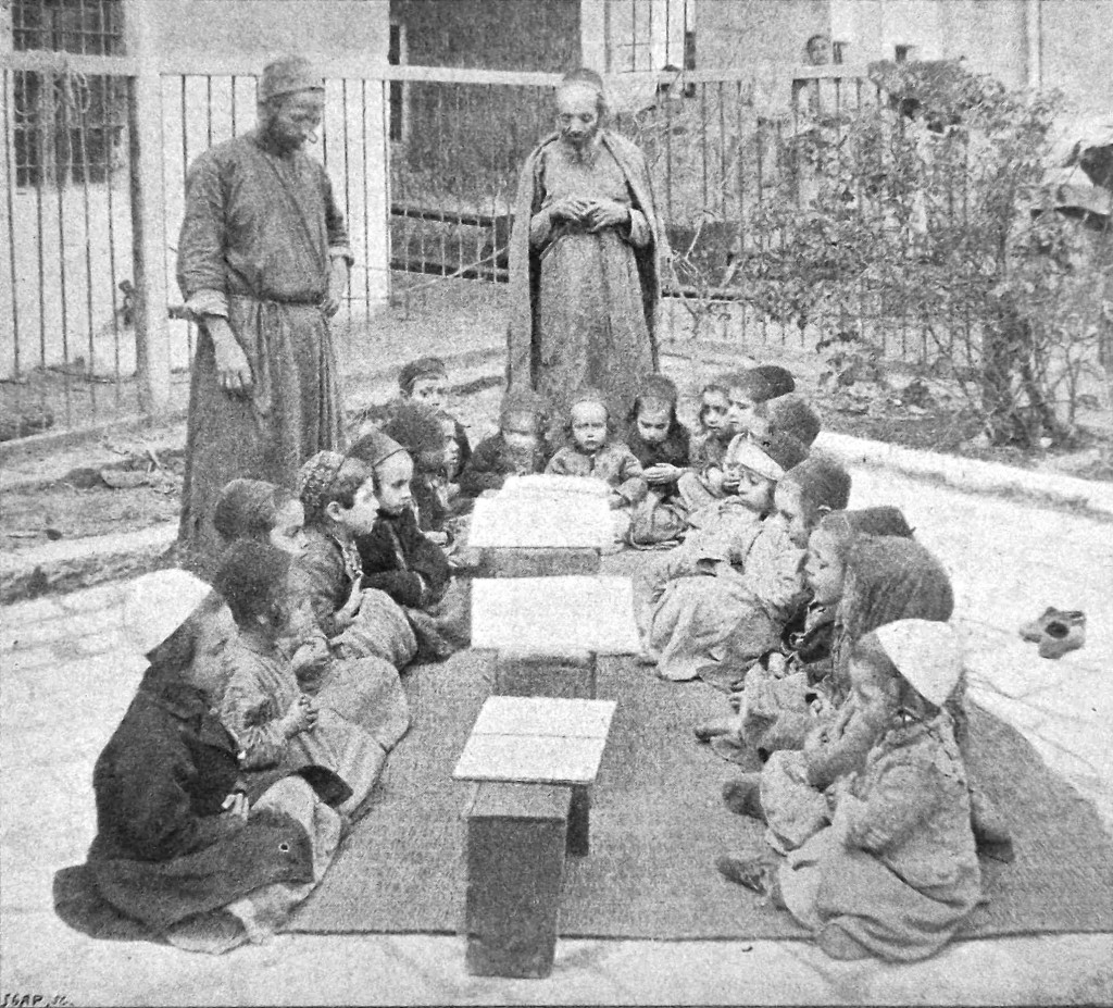 Two rows of boys sitting outdoors on a carpet, facing each other. Male teachers in background.