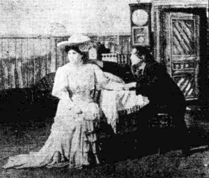 Man and woman seated at a table. He touches her hand and shoulder. She looks away from him. 